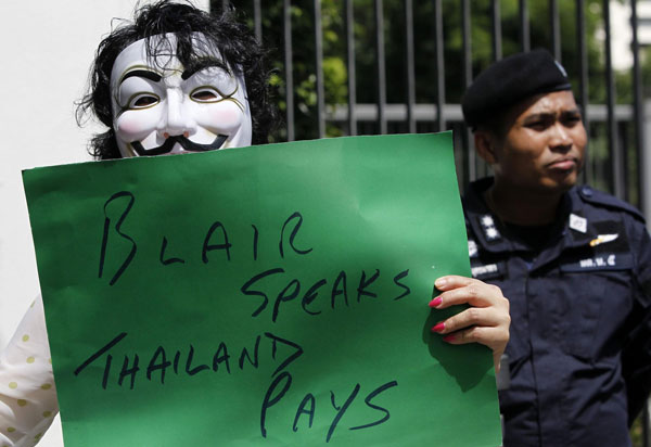 An anti-government protester wearing a Guy Fawkes mask holds a placard as a police officer stands guard outside the British embassy in Bangkok August 27, 2013. Tony Blair's speaker fee draws protest in Thailand